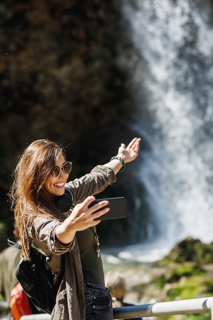 Una giovane donna sorridente che fa selfie con il suo smartphone mentre si gode la vista della bellissima cascata.