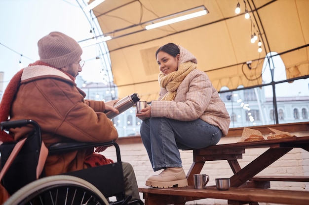 Una giovane donna sorridente con un uomo in sedia a rotelle che condivide bevande calde in inverno