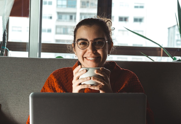 Una giovane donna sorridente davanti al suo laptop nel divano mentre si tiene una tazza di caffè