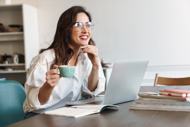 una giovane donna sorridente positiva in posa al chiuso utilizzando il computer portatile che tiene tazza di caffè.