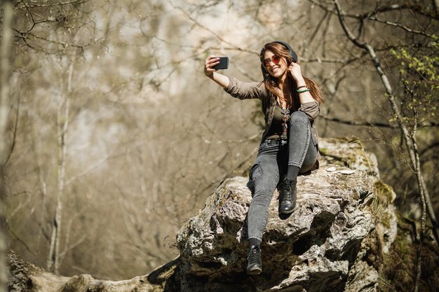Una giovane donna spensierata seduta sulla cima della scogliera della montagna, che fa un selfie e si gode la vita all'aria aperta.