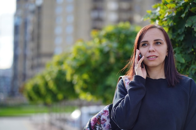 Una giovane donna sta e parla utilizzando uno smartphone sullo sfondo di alberi verdi in una giornata di sole Donna in abiti casual che risponde a una telefonata mentre si trova su sfondo sfocato della strada