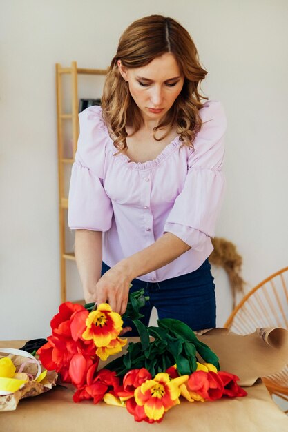 Una giovane donna sta imballando un bouquet festivo in carta da imballaggio Il fiorista fa un assemblaggio con tulipani rossi in officina Una donna al lavoro in una piccola impresa o hobby
