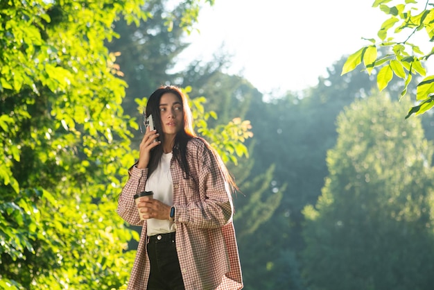 Una giovane donna sta parlando al telefono, beve caffè e si fa un selfie nel parco.