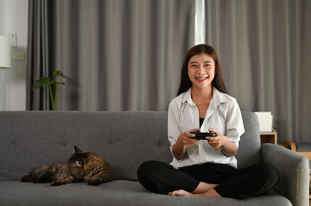 Una giovane donna sta tenendo il joystick e sta giocando ai videogiochi mentre è seduto sul divano di casa.