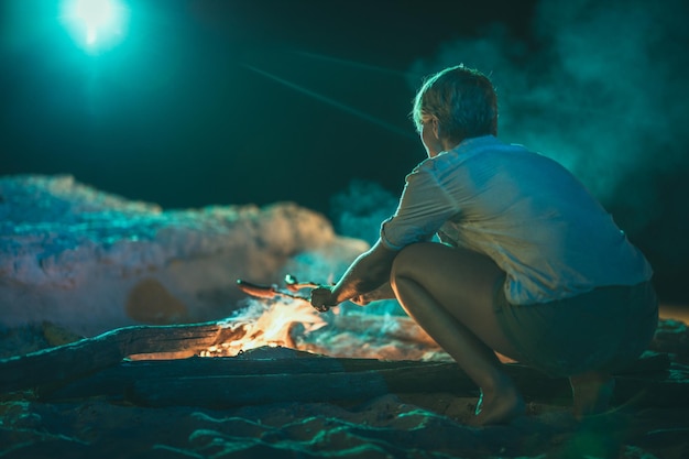 Una giovane donna sta trascorrendo del tempo vicino al fuoco sulla spiaggia sabbiosa la sera. Sta friggendo le salsicce sul bastone.