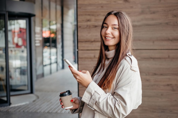 Una giovane donna sta usando un telefono cellulare sorridendo e guardando la telecamera vicino a un edificio per uffici