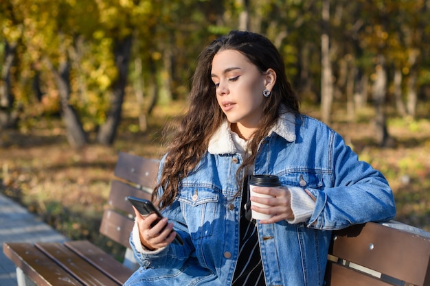 Una giovane donna su una panchina nel parco autunnale con caffè e uno smartphone in mano