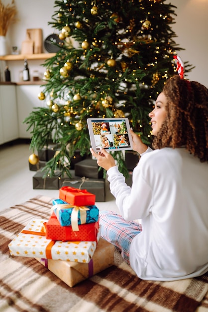 Una giovane donna sullo sfondo di un albero di Natale con regali con un tablet ha una videochiamata.