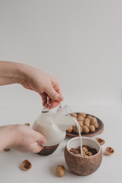 Una giovane donna versa il latte in una ciotola con muesli. Una mano tiene una crema, una brocca di latte. Una sana colazione a base di muesli, farina d'avena, cereali, noci.