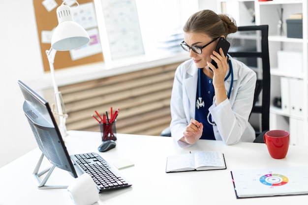 Una giovane donna vestita di bianco è seduta alla scrivania in ufficio, parla al telefono e tiene in mano una penna. Uno stetoscopio le pende al collo.