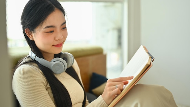 Una giovane e affascinante donna asiatica in abiti casuali che legge un libro in soggiorno.