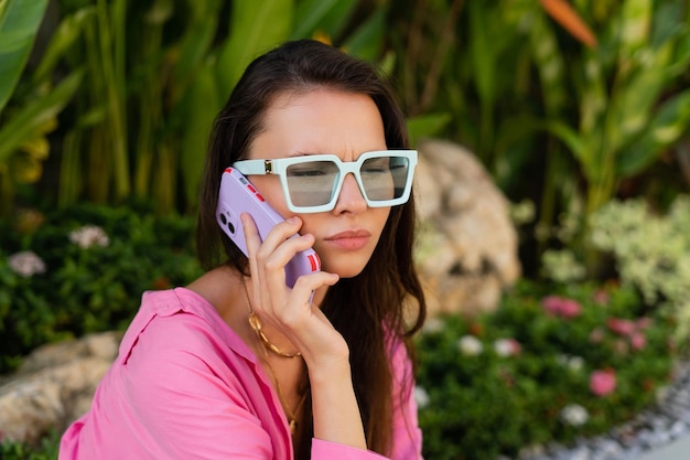 Una giovane e bella donna bruna in occhiali da sole alla moda gioielli collo camicia rosa si siede sullo sfondo di foglie tropicali chiama il telefono è allarmato insoddisfatto indignato