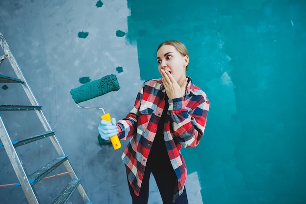 Una giovane e bella donna designer fa riparazioni dipinge un muro grigio con vernice verde con un rullo Camicia a quadri capelli lunghi e jeans