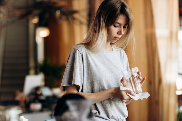 Una giovane e bella ragazza bionda, vestita in stile casual, tiene in mano un bicchiere pulito e lo guarda in una moderna caffetteria. .