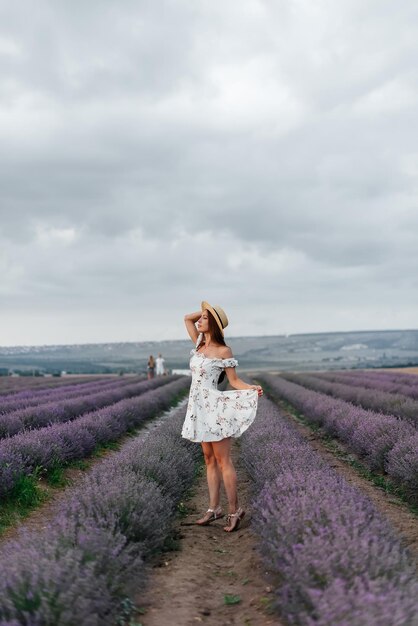 Una giovane e bella ragazza con un vestito e un cappello delicati cammina attraverso un bellissimo campo di lavanda e si gode la fragranza dei fiori Riposo e natura meravigliosa Fioritura della lavanda e raccolta dei fiori