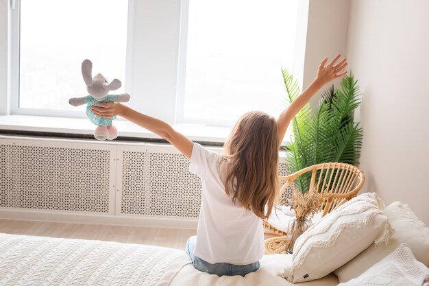 Una giovane e bella ragazza con una maglietta bianca e jeans è seduta sul letto e guarda fuori dalla finestra La ragazza pensa e sogna qualcosa si rallegra al mattino Foto di alta qualità