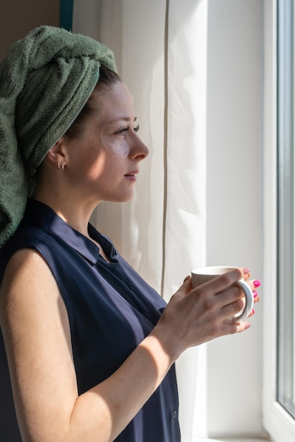 Una giovane e bella ragazza dopo la doccia con un asciugamano in testa sta a casa guarda fuori dalla finestra e tiene una tazza di bevanda calda Il tempo della spa di riposo non è tutt'uno con se stesso