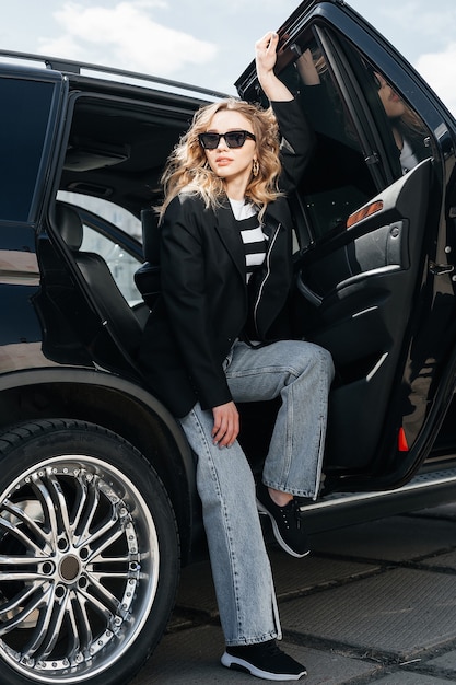 Una giovane e bella ragazza è seduta sul sedile posteriore di un'auto. Una ragazza alla moda in giacca e occhiali esce dall'auto.