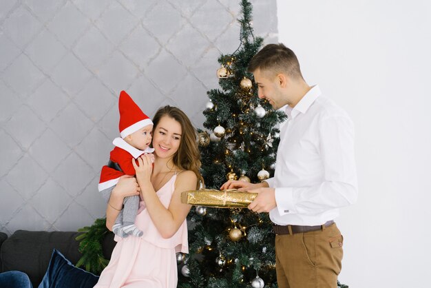 Una giovane famiglia celebra il Natale a casa nel salotto vicino all'albero di Natale. Felice mamma, papà e figlio si godono le vacanze insieme. Padre fa un regalo a mamma e bambino