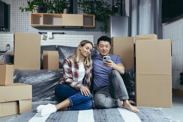 Una giovane famiglia in un nuovo appartamento tra le scatole di cartone seduta sul pavimento e scegliendo felicemente nuovi mobili negozio online utilizza l'app del telefono per lo shopping