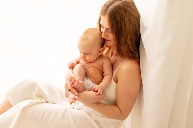 una giovane madre abbraccia dolcemente un bambino tra le braccia mentre è seduta alla finestra