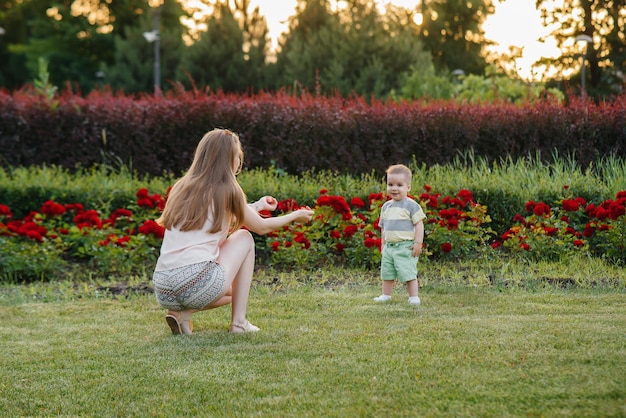 Una giovane madre carina aiuta e insegna al suo piccolo figlio a muovere i primi passi durante il tramonto nel parco sull'erba.