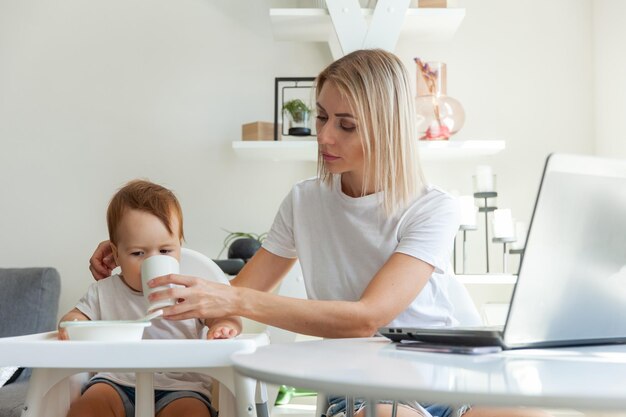 Una giovane madre dà da bere a un bambino piccolo da una tazza mentre è seduta a un tavolo con un portatile Il concetto di cura materna