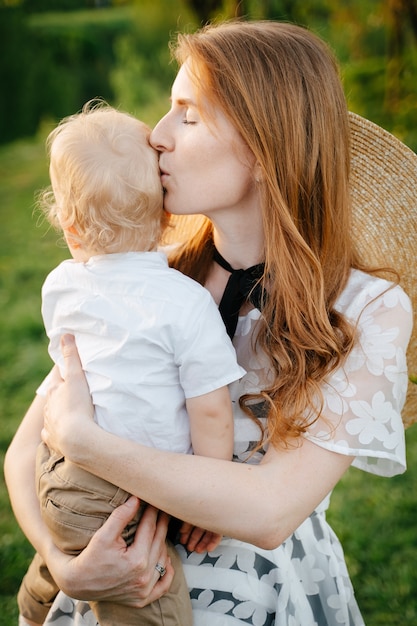 Una giovane madre dai capelli rossi lenisce suo figlio tenendolo tra le braccia e baciandolo sulla testa sullo sfondo della festa della mamma verde
