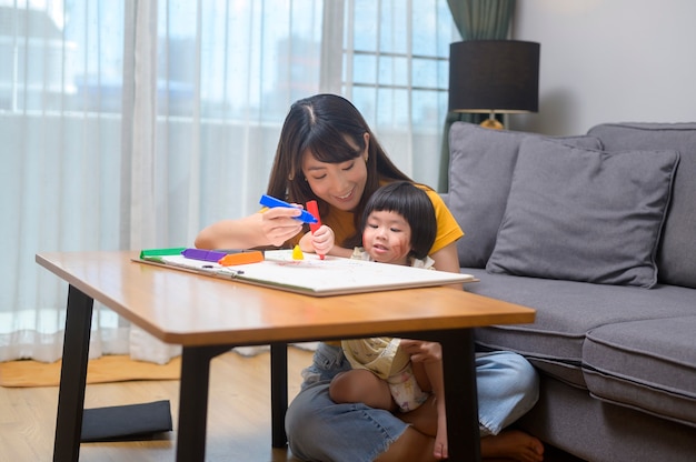 Una giovane mamma che aiuta la figlia a disegnare con matite colorate nel soggiorno di casa.