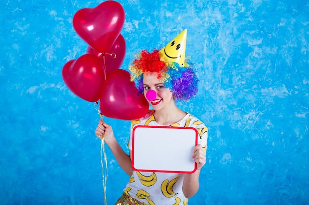Una giovane ragazza allegra in un vestito da clown che posa davanti a una telecamera su uno sfondo blu 1 aprile concetto di Giorno dei pazzi compleanno