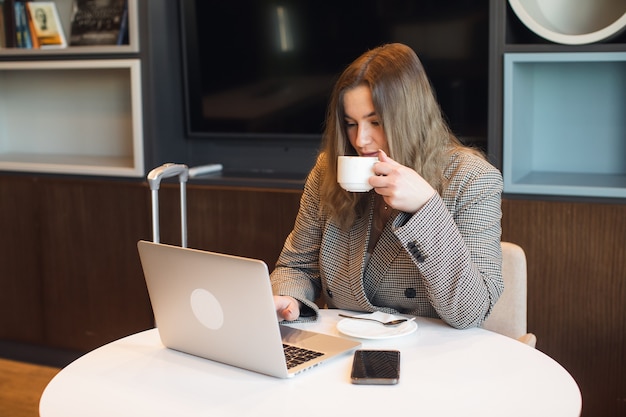 Una giovane ragazza attraente e grassoccia è seduta in un bar a bere caffè e lavorare su una foto di alta qualità del computer portatile