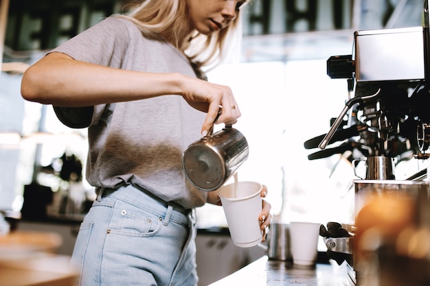 Una giovane ragazza bionda magra, che indossa abiti casual, viene mostrata mentre aggiunge latte al caffè in un'accogliente caffetteria. .