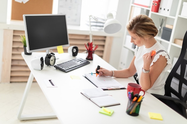 Una giovane ragazza bionda si siede alla scrivania di un computer in ufficio, tiene in mano un pennarello arancione e lavora con i documenti.