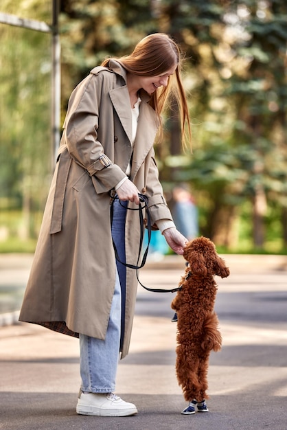 Una giovane ragazza bionda sorridente in un elegante impermeabile beige e jeans gioca con il suo whil barboncino giocattolo