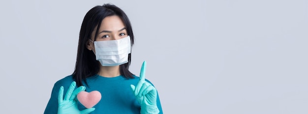 Una giovane ragazza bruna con una maschera medica protettiva tiene in mano un cuore rosa che simboleggia la vita