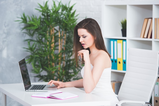 Una giovane ragazza bruna in un abito bianco è seduta in ufficio al tavolo. La donna lavora al computer portatile.