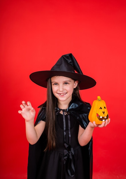 Una giovane ragazza bruna in un costume da strega con un cappello tiene una zucca su uno sfondo rosso con una copia dello spazio