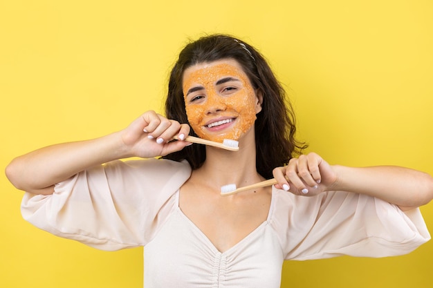 Una giovane ragazza carina con una maschera scrub sul viso si lava i denti contro uno sfondo giallo