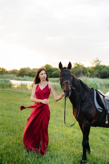 Una giovane ragazza carina in un vestito rosso posa in un ranch con uno stallone purosangue al tramonto. Amore e cura per gli animali.