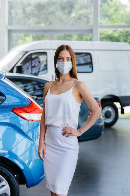 Una giovane ragazza carina ispeziona una nuova auto in una concessionaria di automobili in una maschera durante la pandemia. La compravendita di autoveicoli, nel periodo della pandemia.