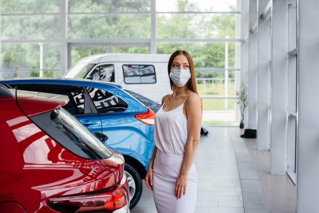 Una giovane ragazza carina ispeziona una nuova auto presso un concessionario di auto in una maschera durante la pandemia. La vendita e l'acquisto di automobili, nel periodo di pandemia.