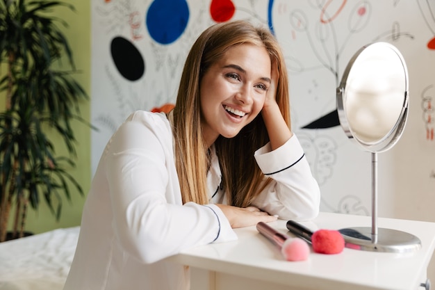 una giovane ragazza carina sorridente ottimista positiva in pigiama a casa vicino allo specchio con pennelli per il trucco.