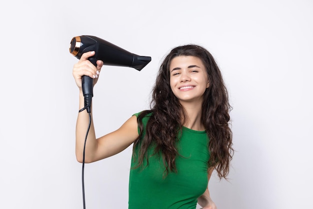 Una giovane ragazza carina vestita con un top verde asciuga i suoi bei capelli lunghi e setosi con un asciugacapelli su uno sfondo bianco