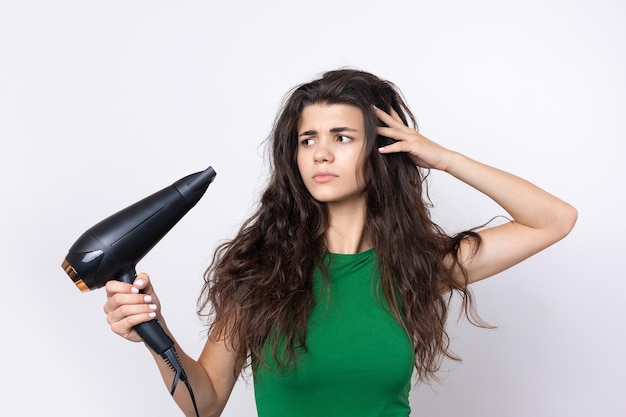 Una giovane ragazza carina vestita con un top verde asciuga i suoi bei capelli lunghi e setosi con un asciugacapelli su uno sfondo bianco