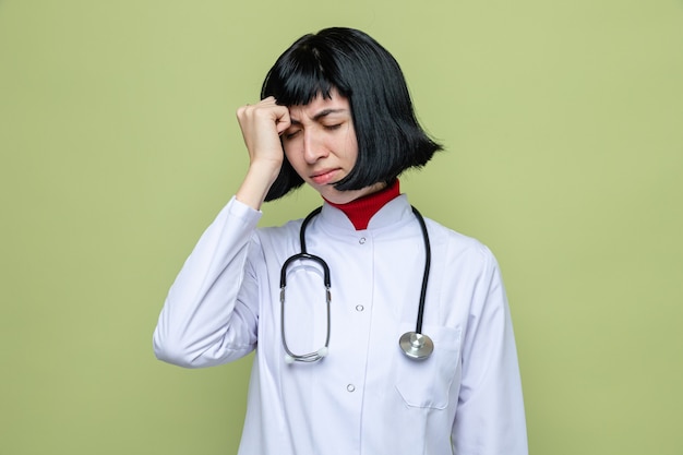 Una giovane ragazza caucasica dolorante in uniforme da medico con stetoscopio mette la mano sulla fronte