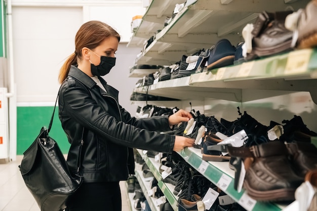 Una giovane ragazza caucasica in una maschera medica nera sceglie vestiti, prodotti di scarpe al supermercato. Il concetto di distanza sociale e