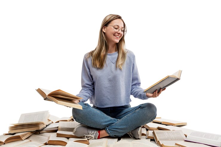 Una giovane ragazza che ride in jeans e una maglietta blu siede su una pila di libri aperti e tiene i libri in mano. Istruzione e conoscenza. .