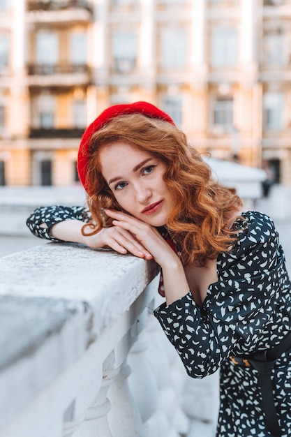 Una giovane ragazza con i capelli rossi in un vestito nero e un berretto rosso sullo sfondo della città