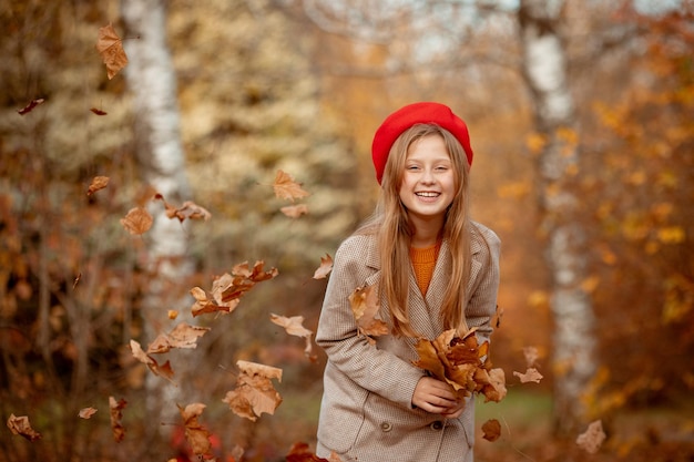 Una giovane ragazza con un berretto rosso e un cappotto tiene in mano un mazzo di foglie autunnali esulta in autunno